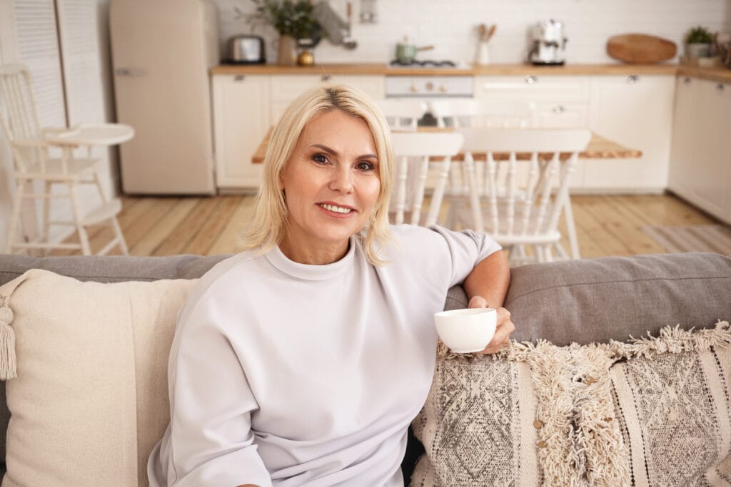 Portrait of attractive middle aged european female with blonde hair relaxing indoors with cup of coffee. beautiful 45 year old woman sitting on couch in living room, enjoying tea, smiling at camera