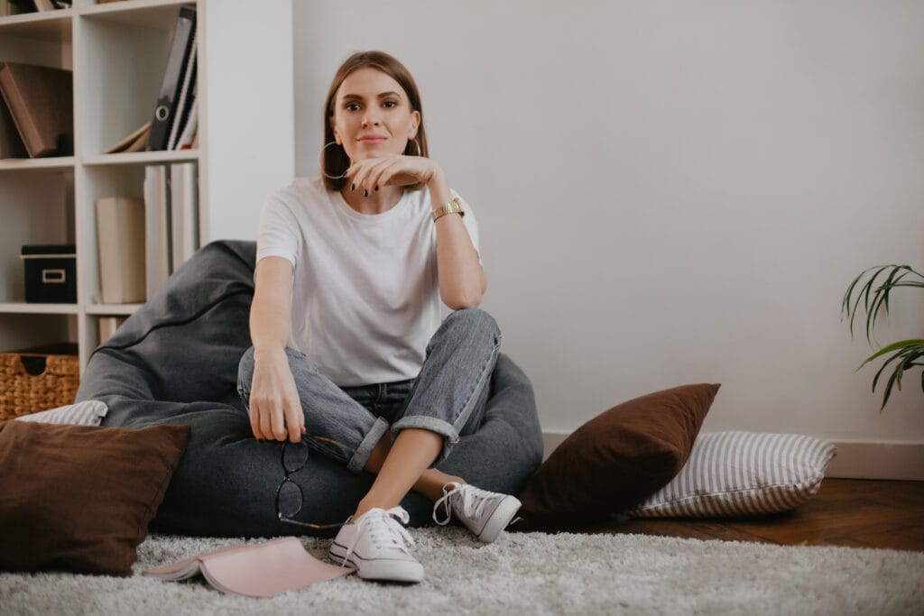 Snapshot of calm young woman in stylish outfit sitting in gray bag chair in white room.
